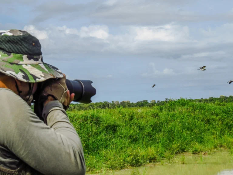 A birdwatcher wearing a camouflage hat and gloves, using a telephoto camera lens to capture birds flying over a lush green wetland under a partly cloudy sky.